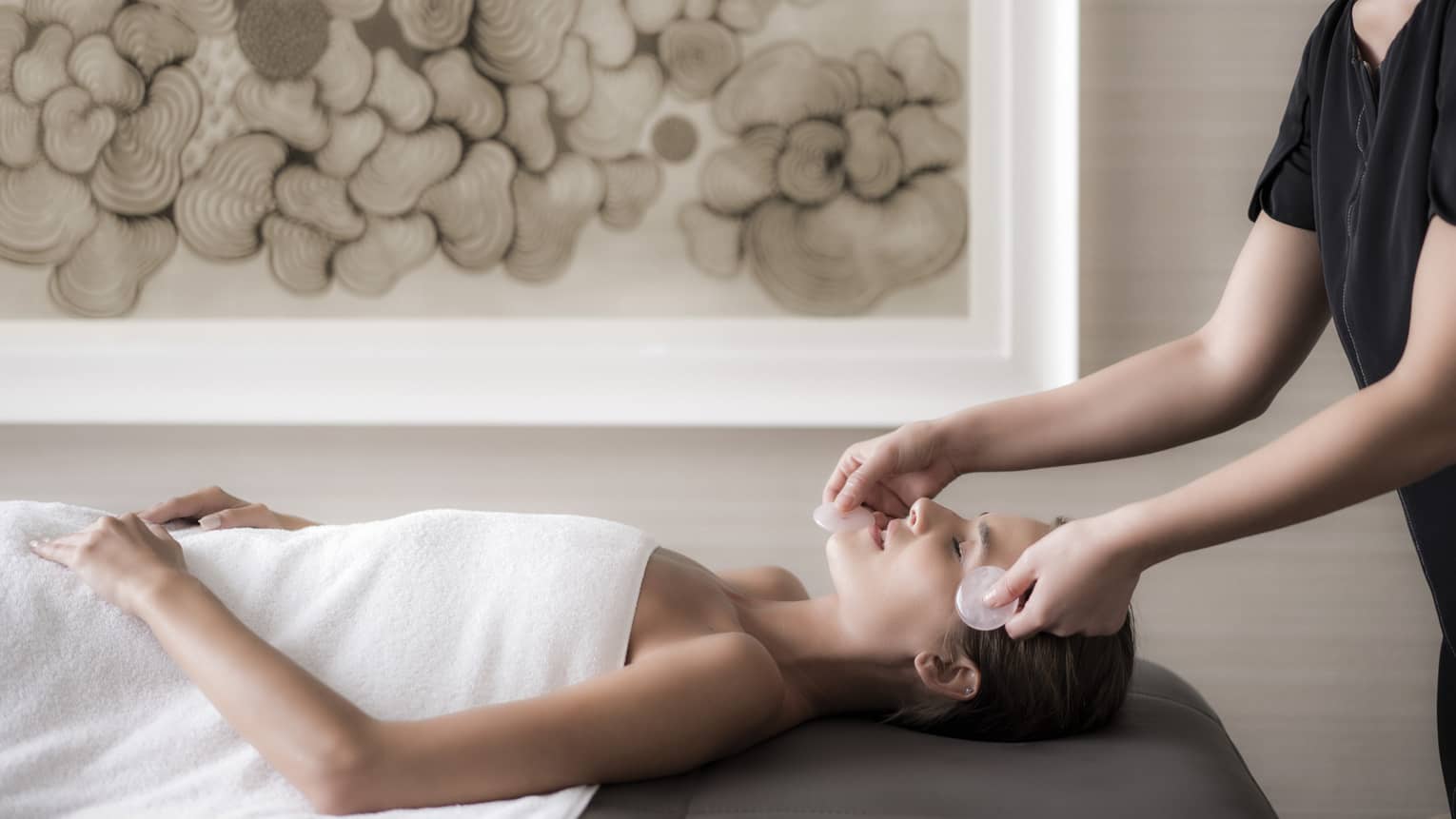 Woman lies on massage table wearing white towel as spa attendant massages her face with stones