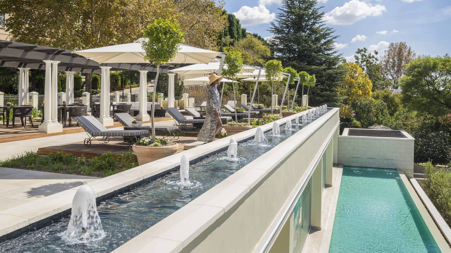 Woman in flowing dress by lounge patio chairs, long water fountain by swimming pool