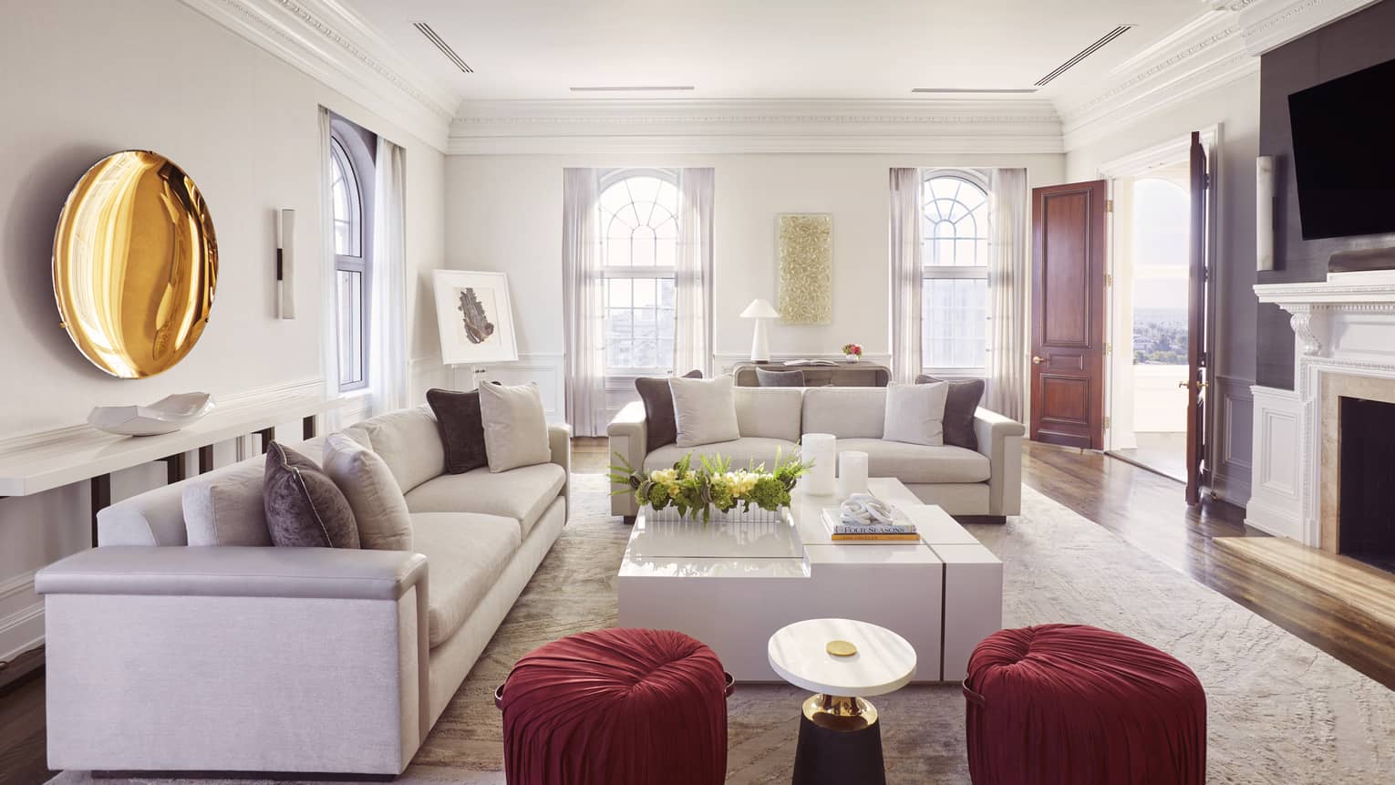 Living room with two sofas, large white square coffee table, two red cushioned stools, gold plate artwork on wall, TV and fireplace
