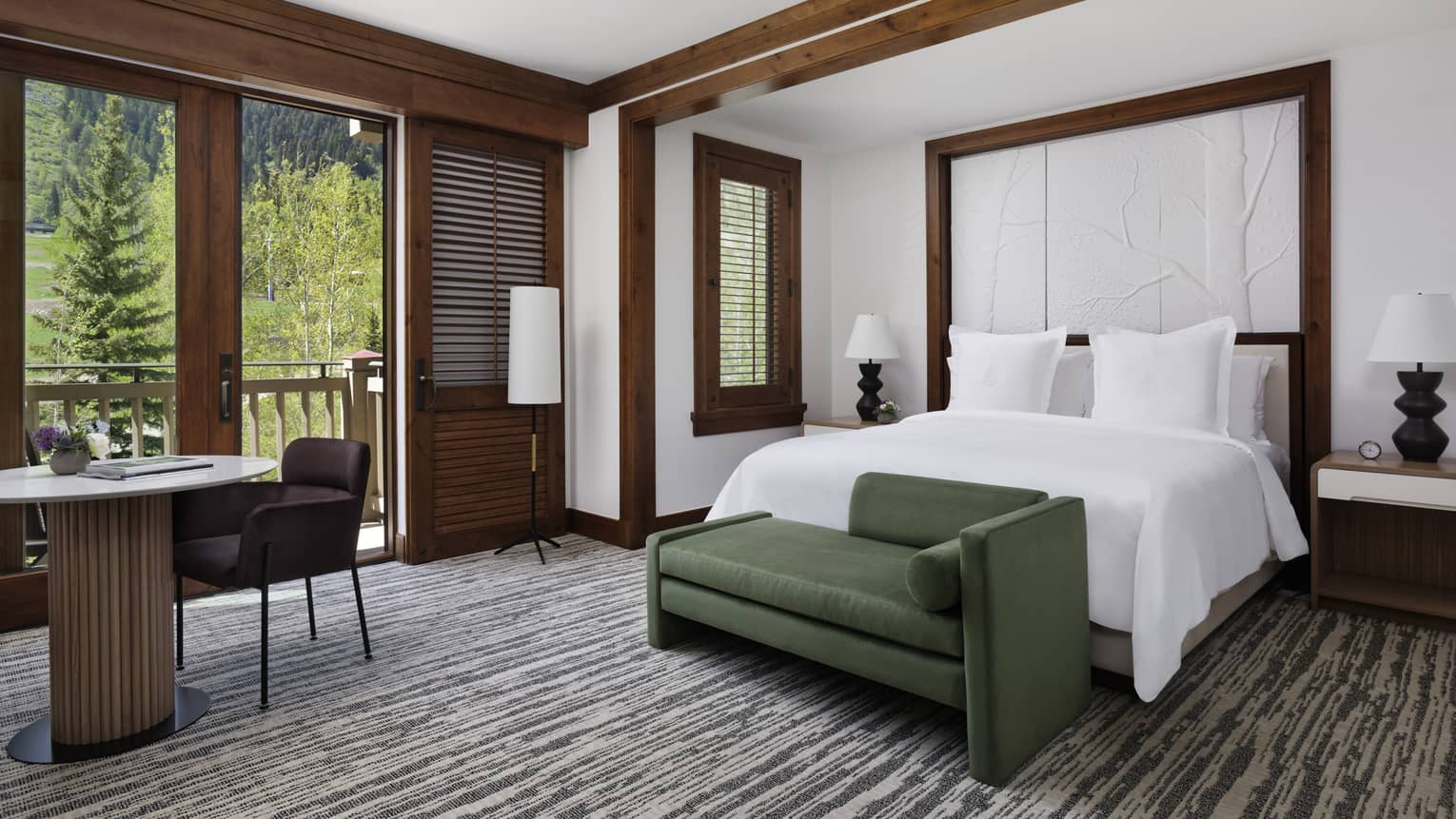 Hotel room with king bed, green chaise, table and two chairs, and balcony view of forest