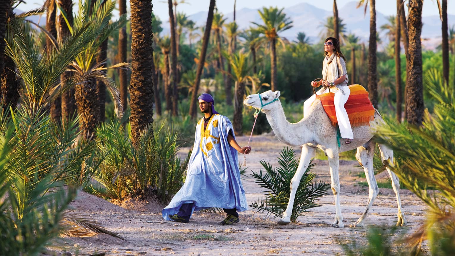 Man leads woman riding camel down sandy path surrounded by palm trees
