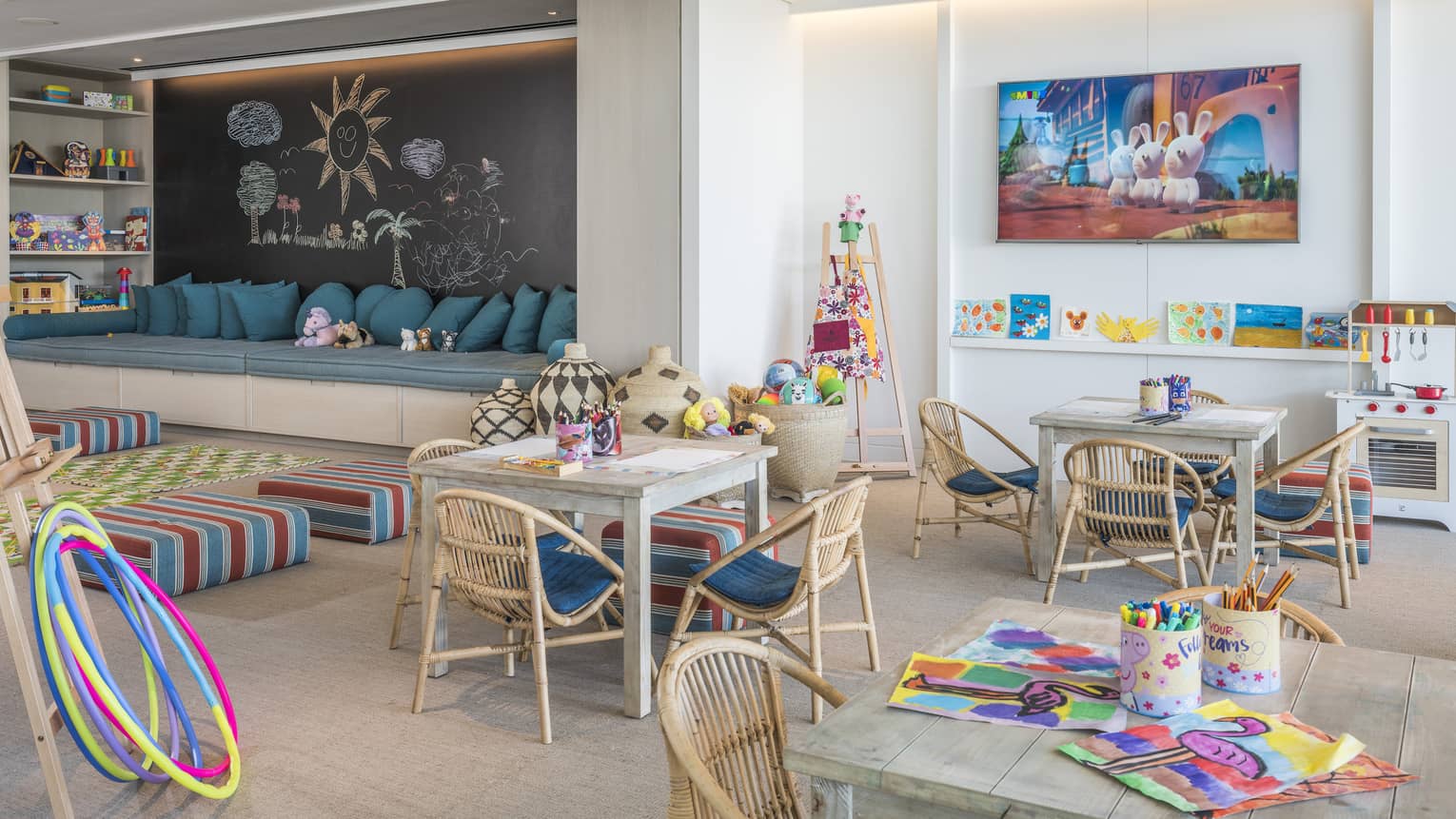 A well-lit playroom with wooden tables and chairs, colourful floor cushions and toys neatly placed on shelves.