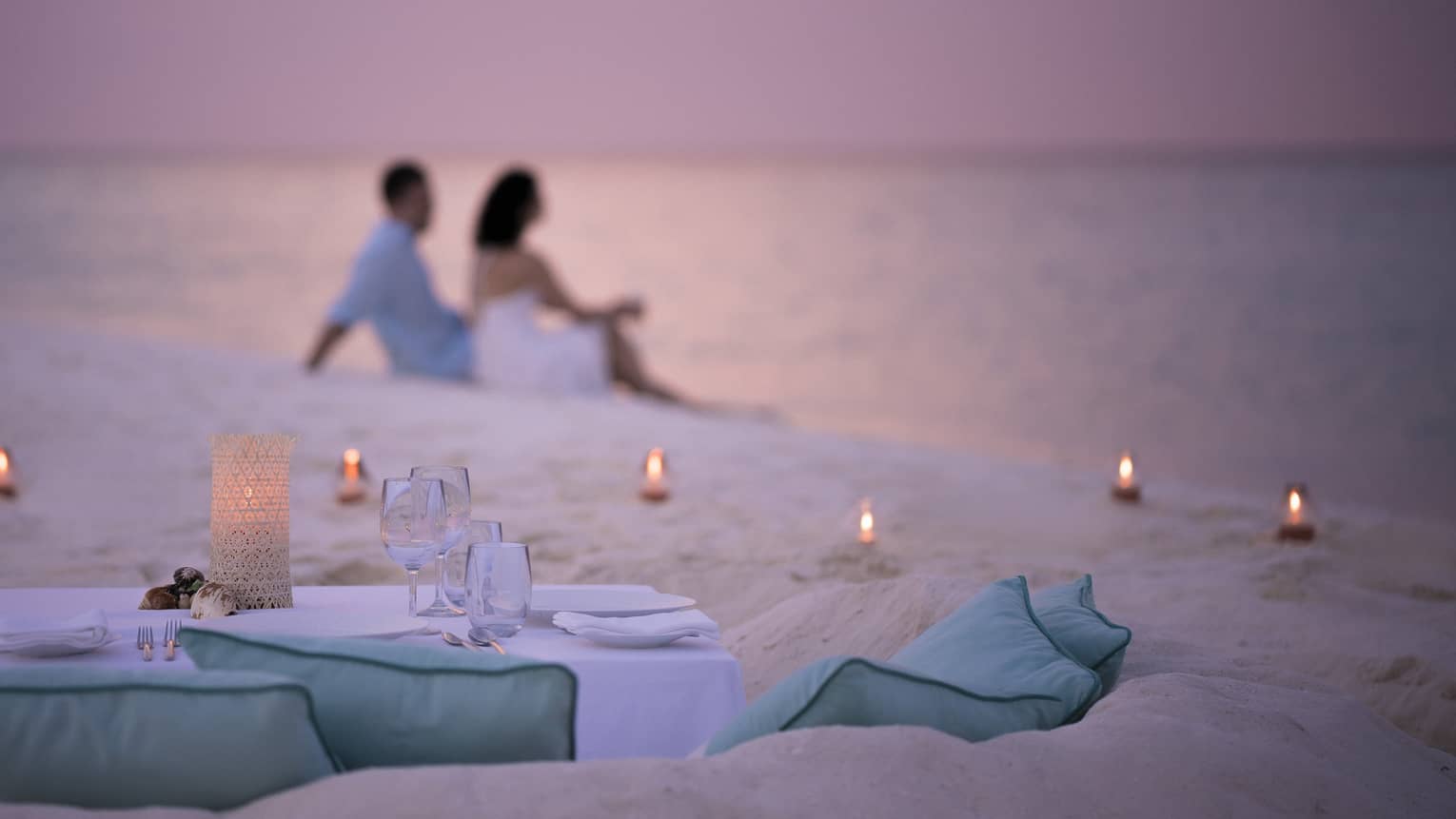 Private dining table in front of sand beach with tealight candles, couple sitting in sand