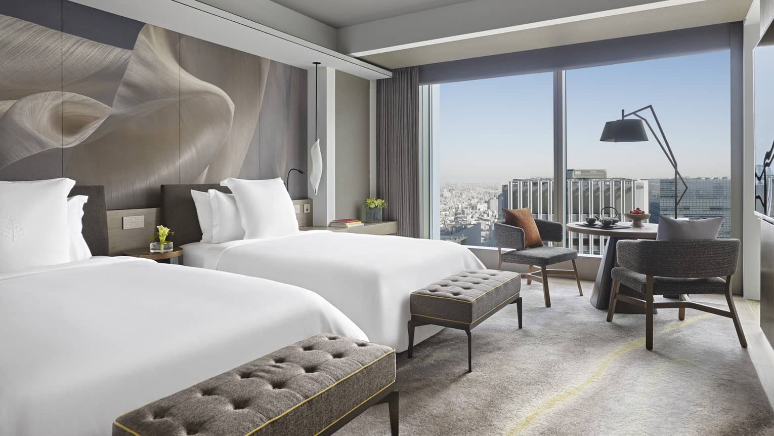 Guest room with two double beds, upholstered benches, a seating area and floor-to-ceiling windows offering city view