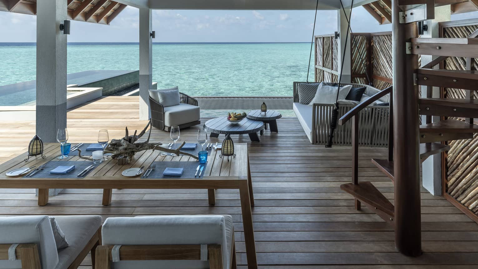 Outdoor terrace of an over-water villa, featuring wooden floors, table and chairs, lounge seating, staircase