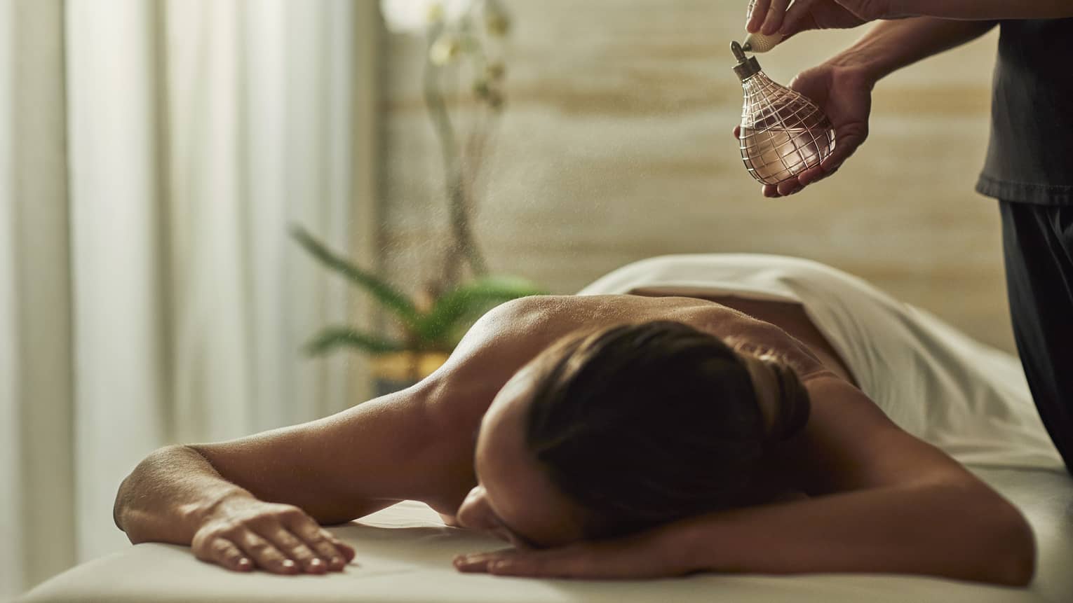 Spa staff sprays perfume bottle over woman lying on massage table