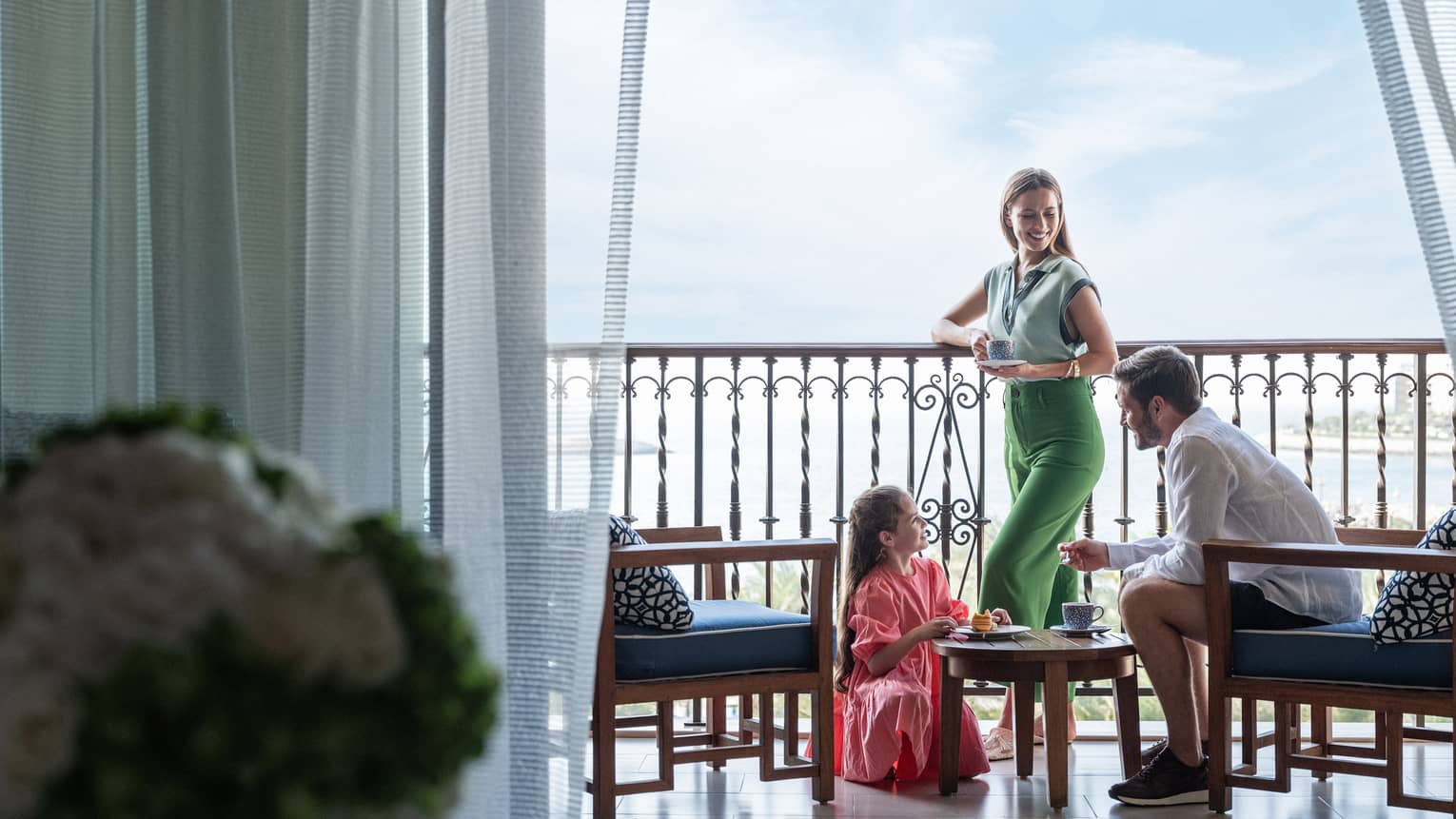 A couple and their daughter stand on a balcony below a bright blue sky.
