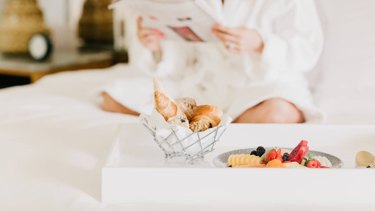 Pastries, fruit on in room dining tray, woman wearing white bath robe reads in background