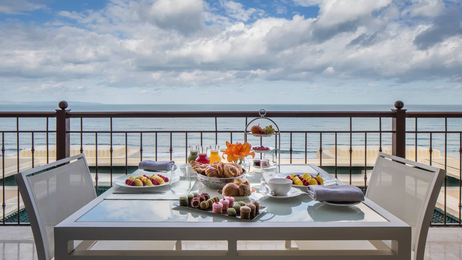 Balcony table set with fresh pastries, cakes, fruits, sea in background
