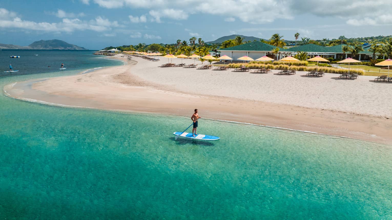 Man stand-up paddleboarding near a shoreline lined with a row of yellow beach umbrellas