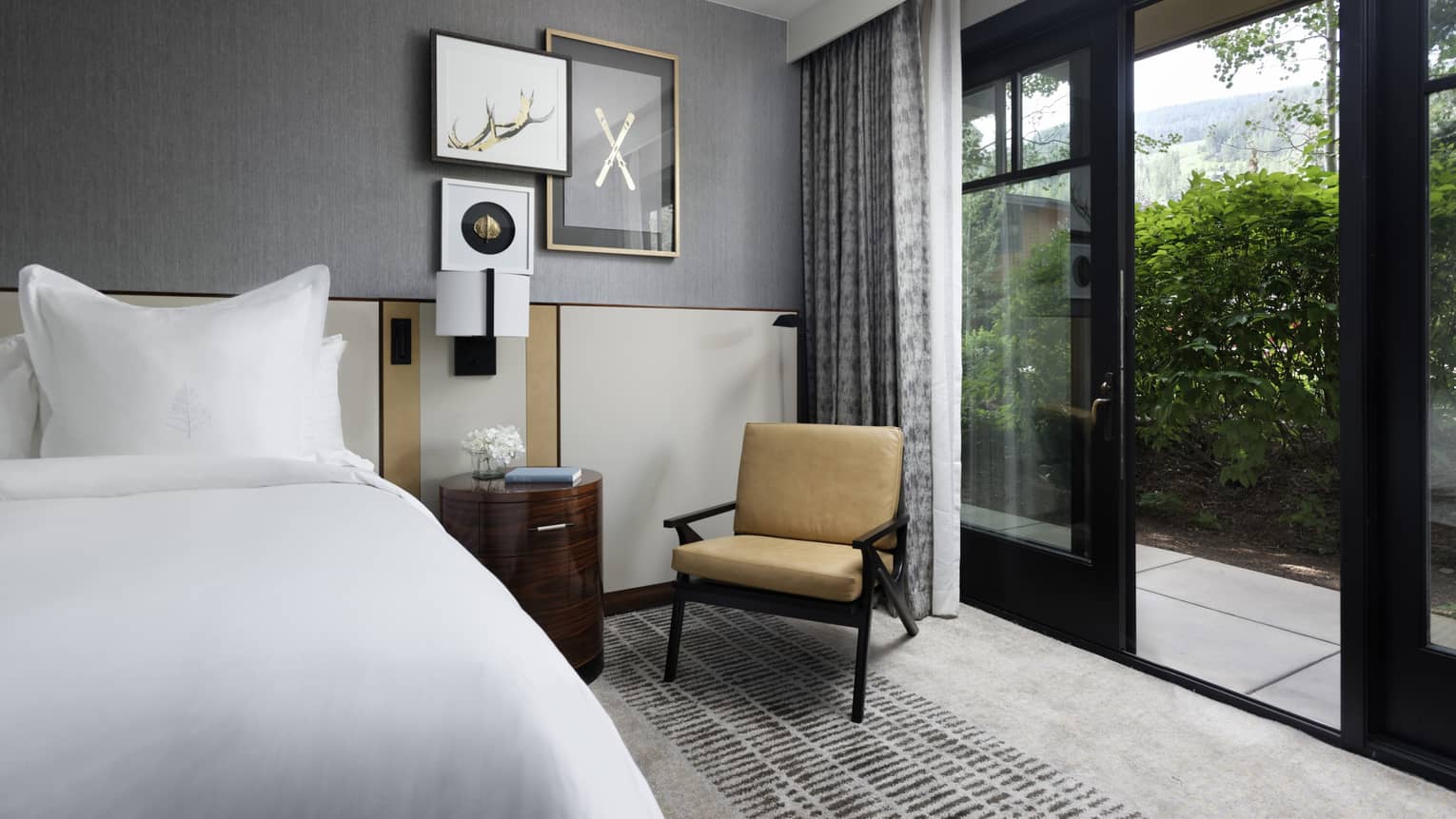 Guest room with bed, white linens, grey walls, wooden side table, lounge chair, sliding glass doors leading to terrace