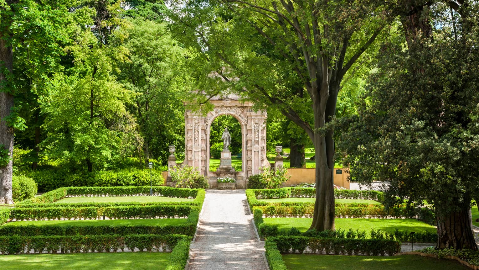 Garden with shrubs, trees in front of stone arch, statue