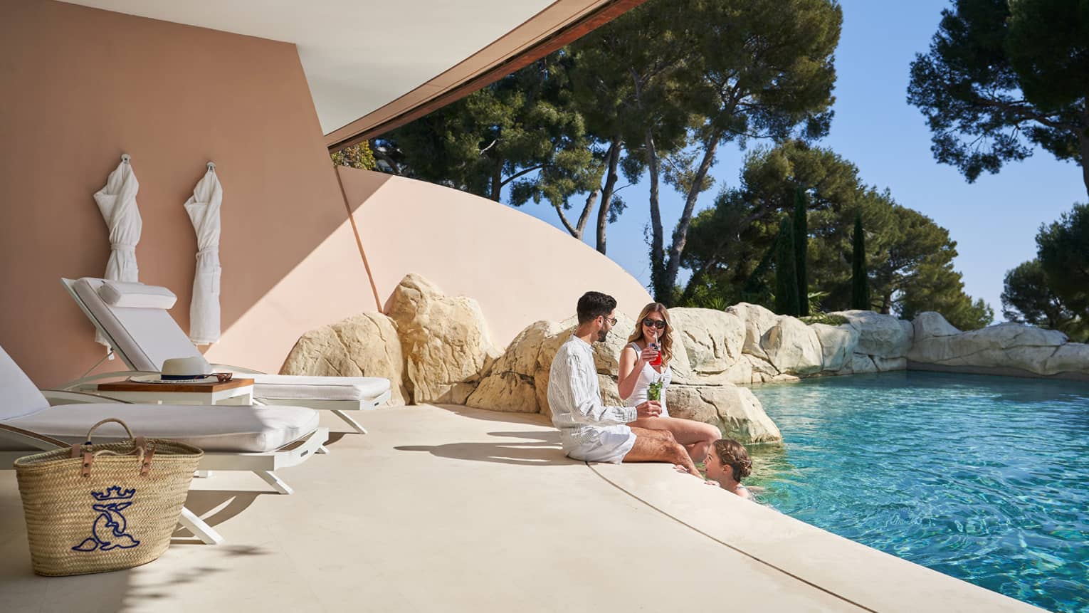 A family lounges on a terrace by an outdoor pool, surrounded by lush green trees