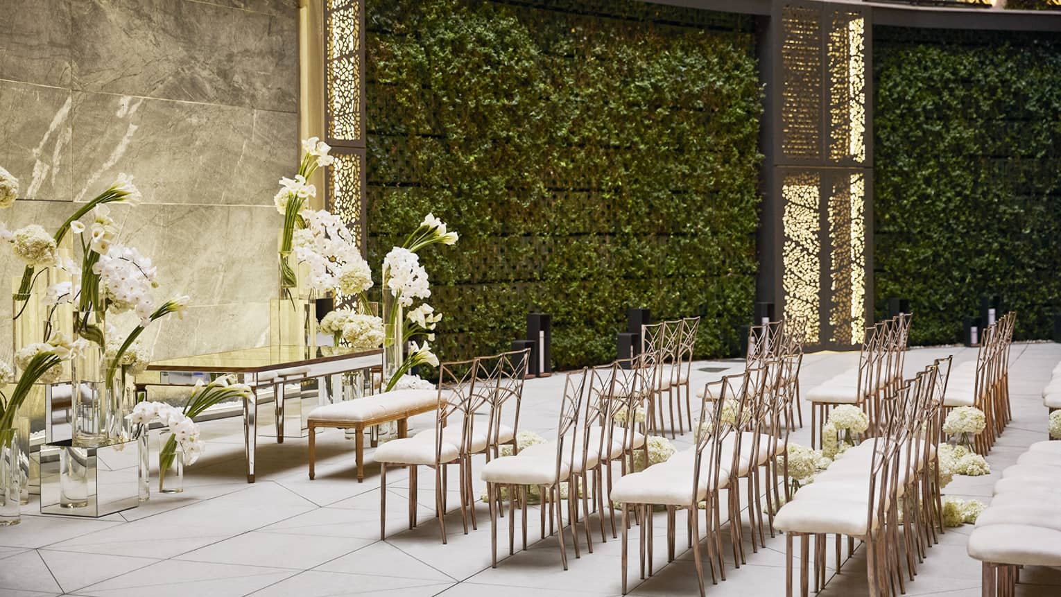 Five rows of white-and-gold chairs set up facing an antique table and bench surrounded by large white floral arrangements, all set in an intertior courtyard featuring curved, vine-filled walls