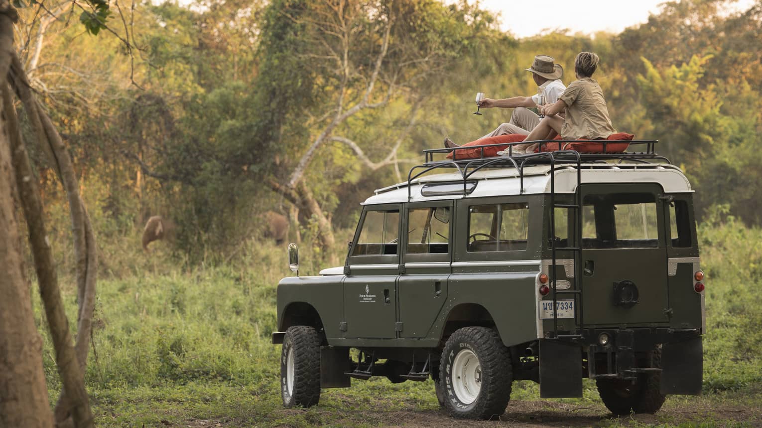 A couple sits on the roof of a Land Rover in the Bush, drinking a cocktail