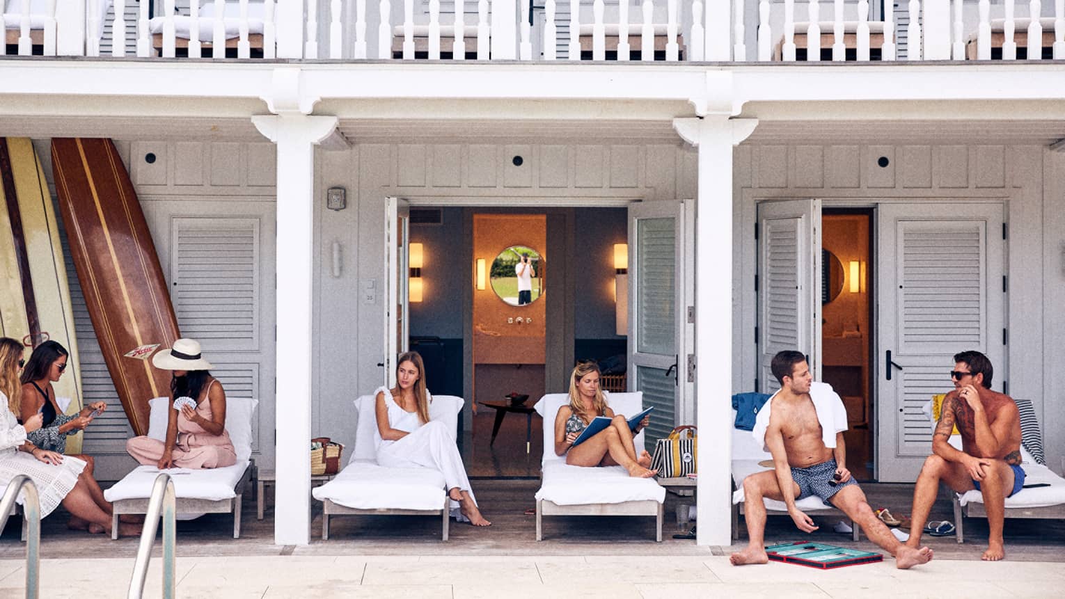 Group of six men and women in bathings suits and leisurewear socializing poolside on lounge chairs