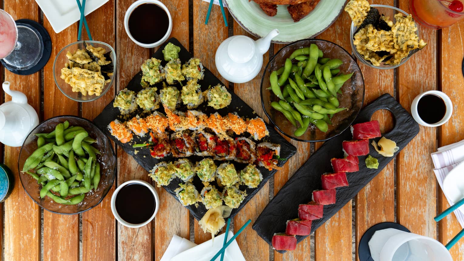 Overhead view of large sushi platter, another platter with one sushi roll, two small bowls of edamame and two small bowls of tempura along with various beverages on a wooden table