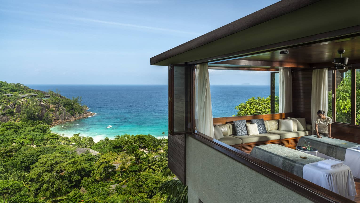 Woman smoothes sheet on side-by-side massage tables in spa room high above trees, ocean
