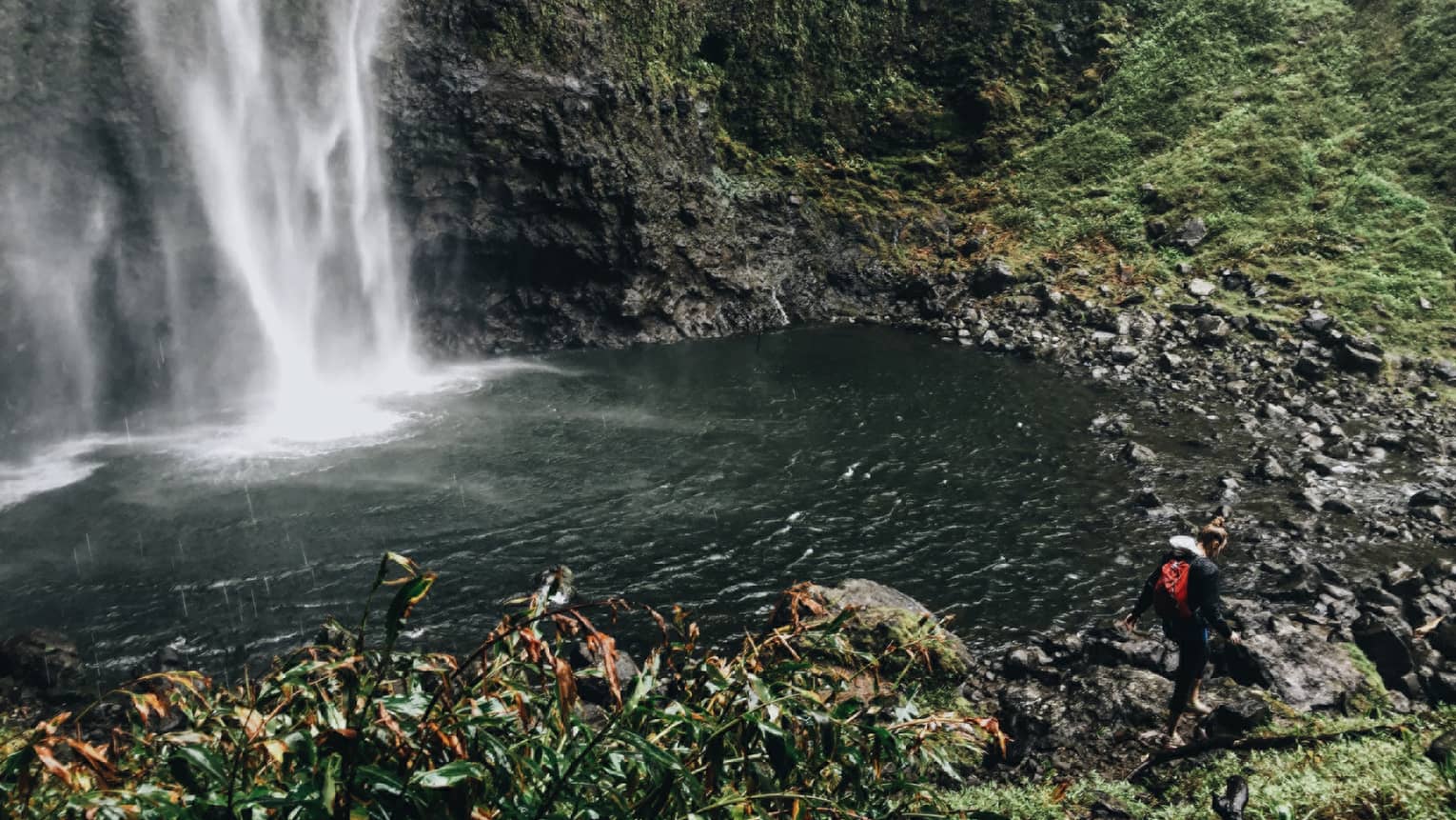 Back of woman hiking over black rocks under tall waterfall, small pond