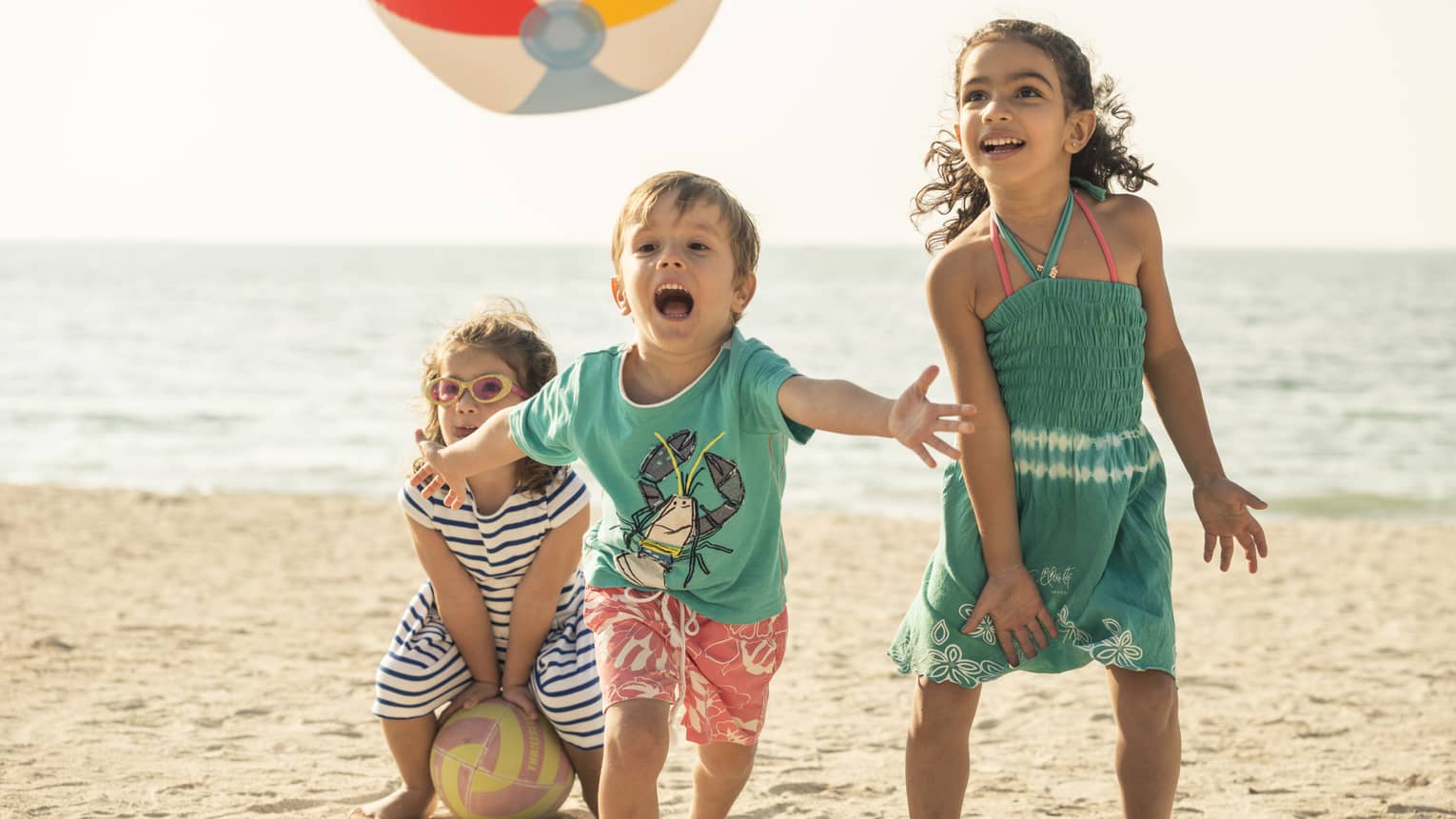 Close-up of three children playing with a beach ball on a sandy beach, the calm ocean and clear sky spanning behind them.