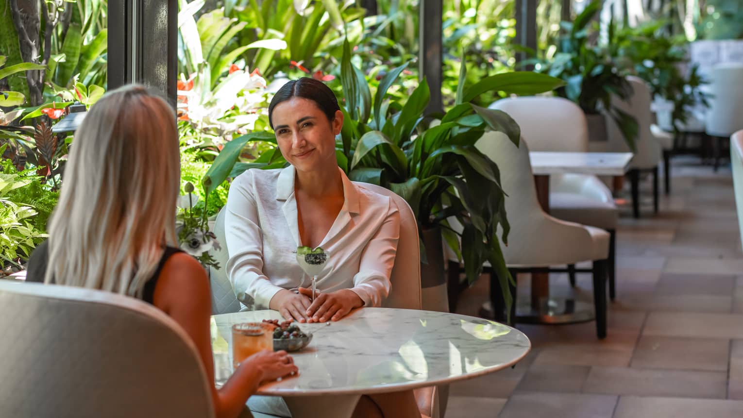 Two women sit at round marble table in indoor-outdoor hotel lounge with cocktails