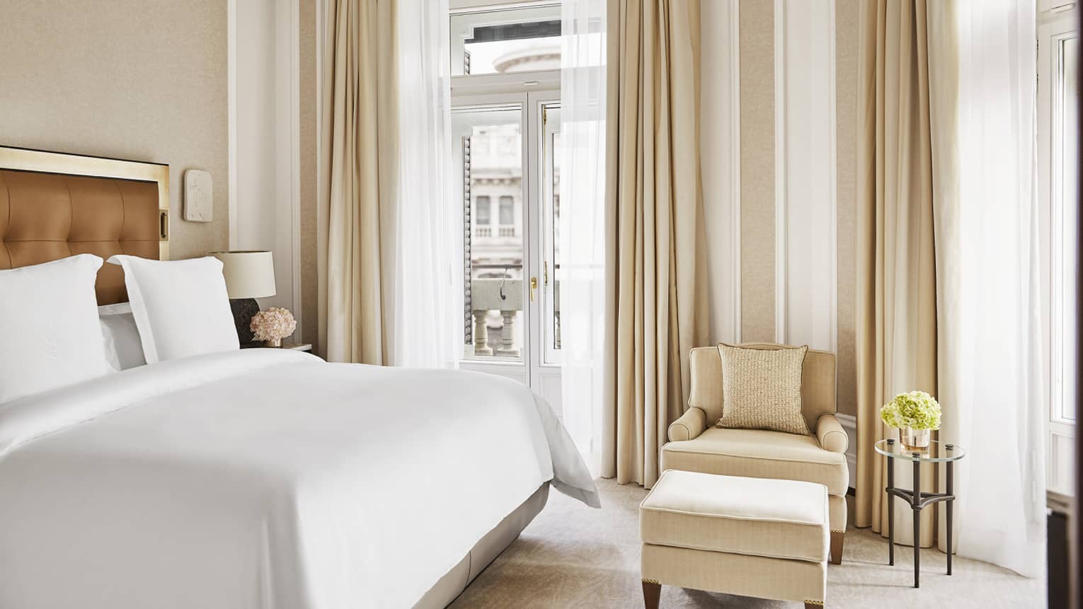 Luxury hotel bedroom with a king bed with a tufted headboard, a cosy armchair with a matching ottoman and a small glass side table with a floral arrangement