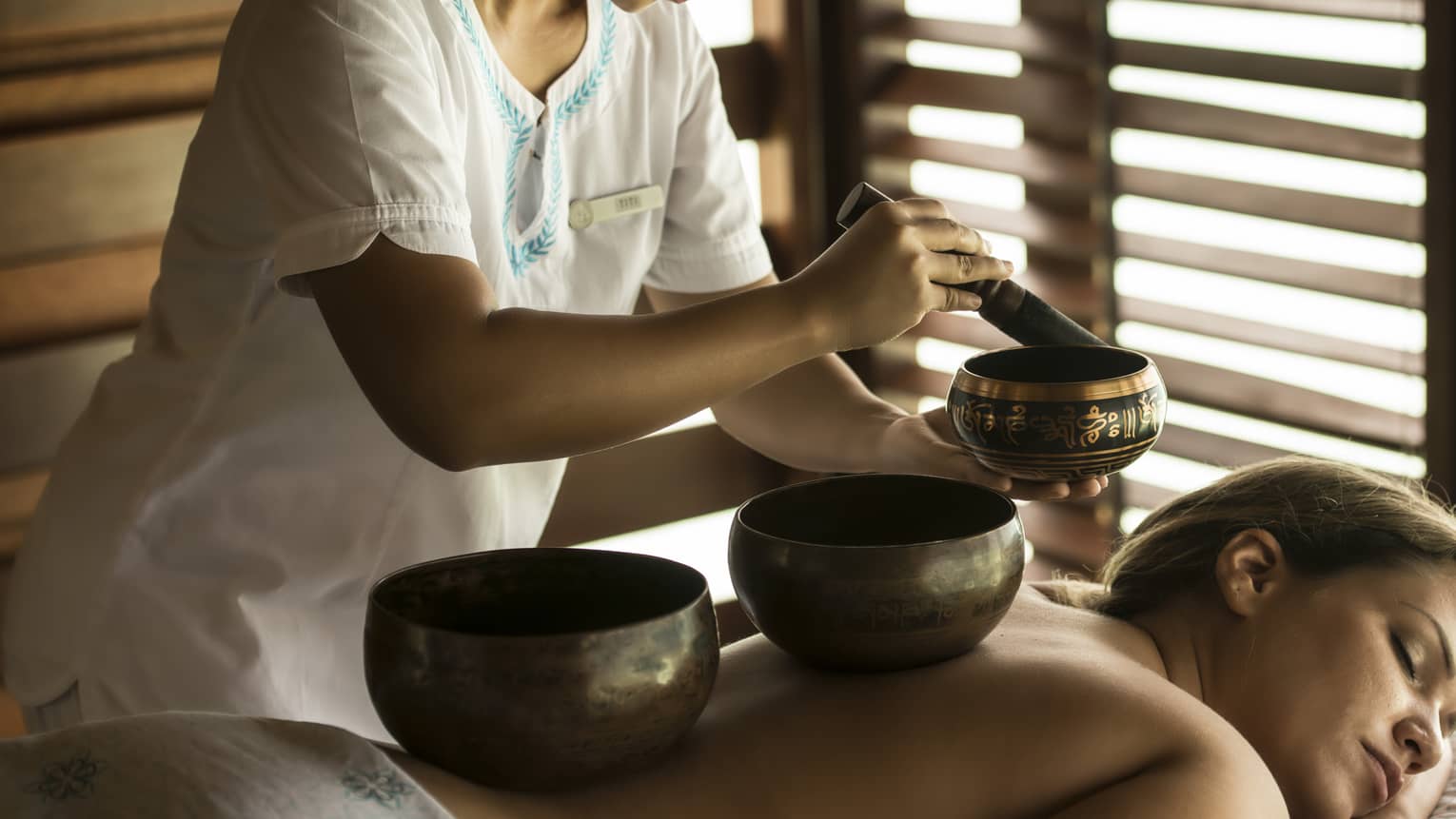 Guest with singing bowls on bare back lies on treatment table which sound specialist plays another singing bowl during a sound healing session