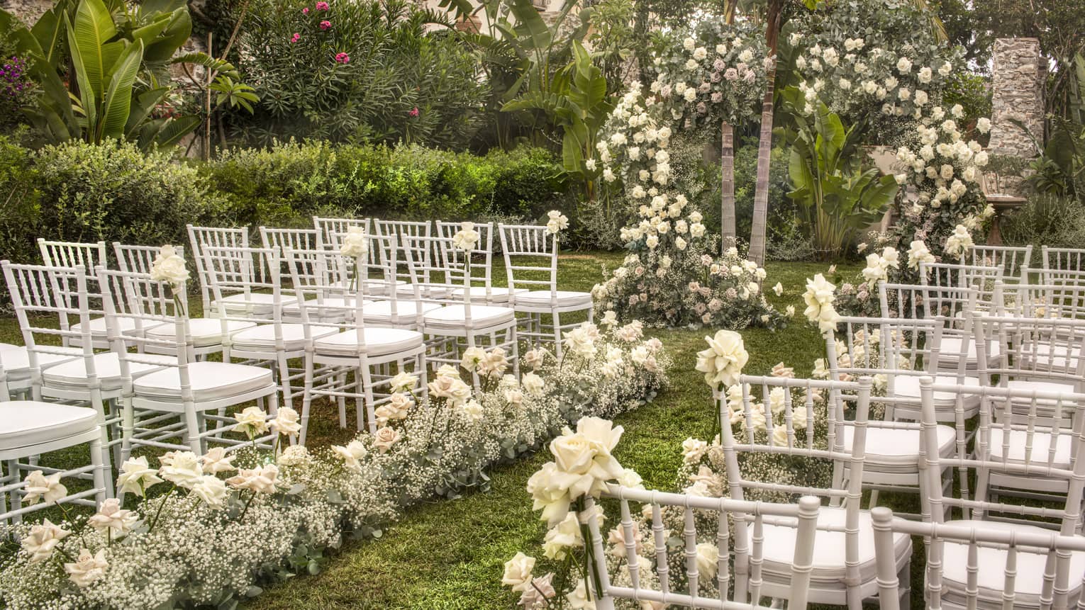 A garden wedding setup featuring rows of white chairs along a floral aisle leading to a flower arch