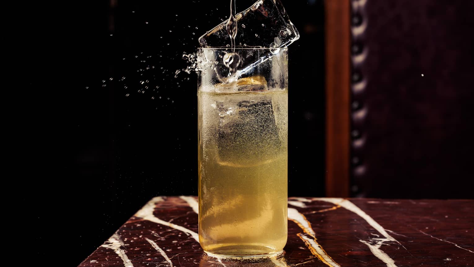 A large rectangular ice cube splashes down into a high-ball glass filled almost to the top with a gold-colored beverage
