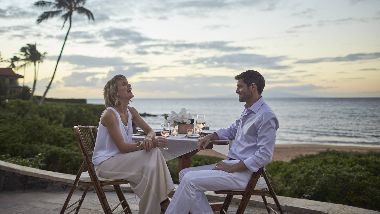Two people enjoying a beachfront dinner at sunset with wine and ocean view