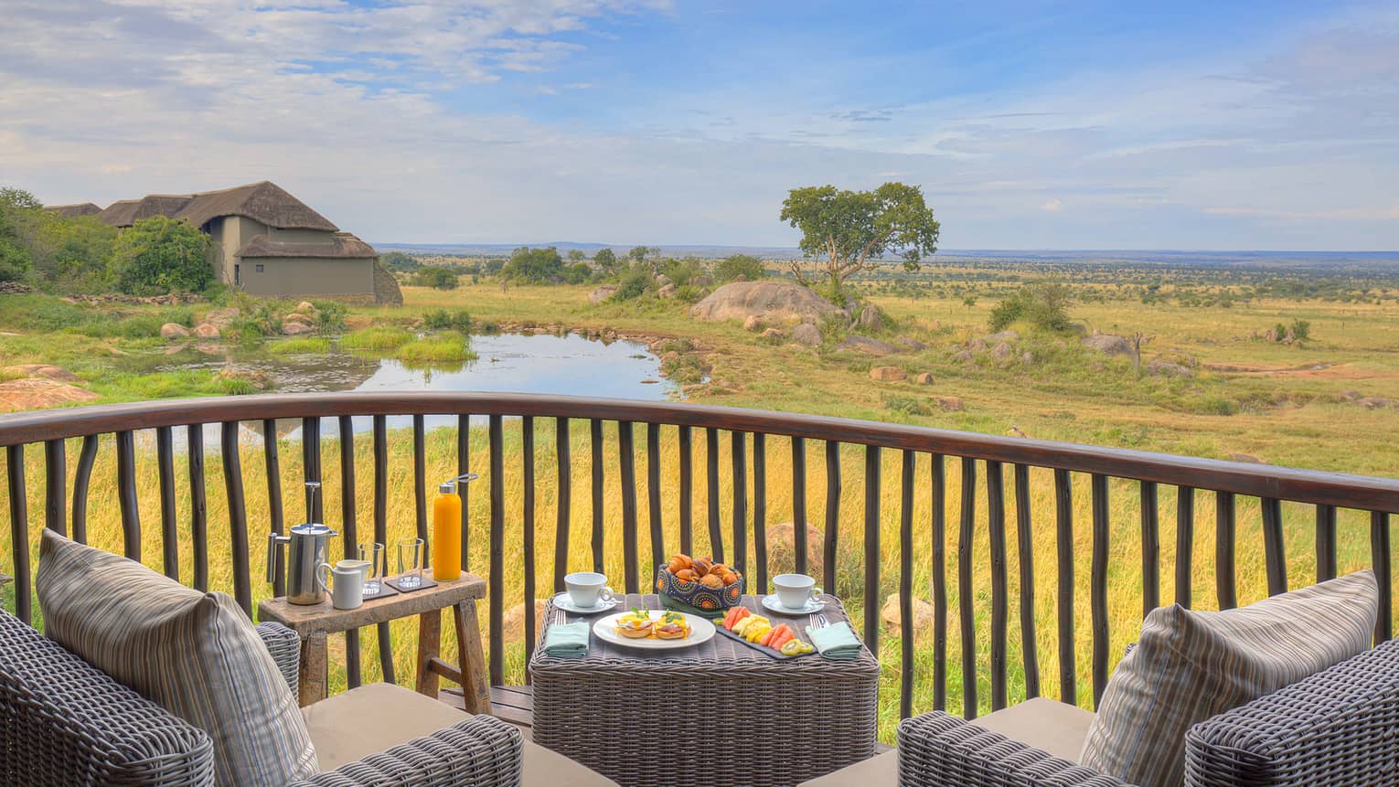 Suite terrace with wicker seating and breakfast setup on square table looking out to watering hole and expansive plains view through rustic vertical wood railing