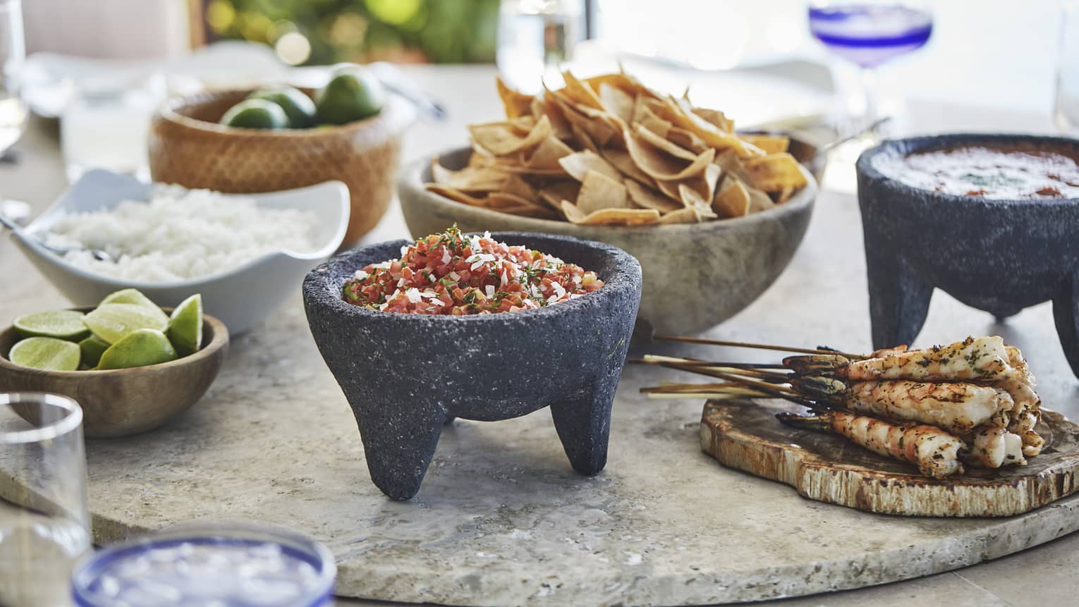 Fresh salsa in lava stone dish near bowl with nacho chips, chicken skewers, limes