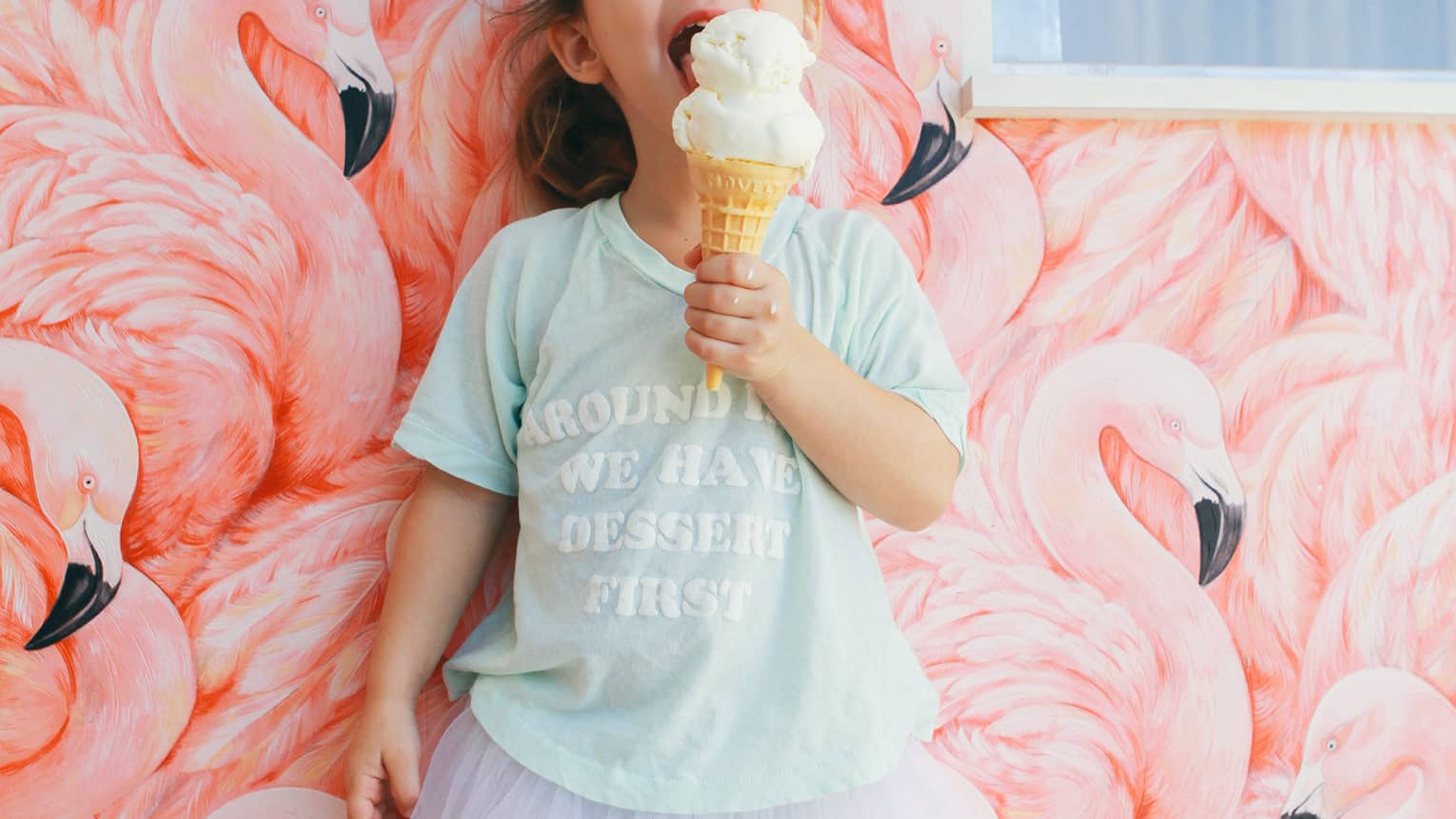 Child in front of a flamingo-motif wall with ice cream cone in one hand, wearing a tutu and t-shirt