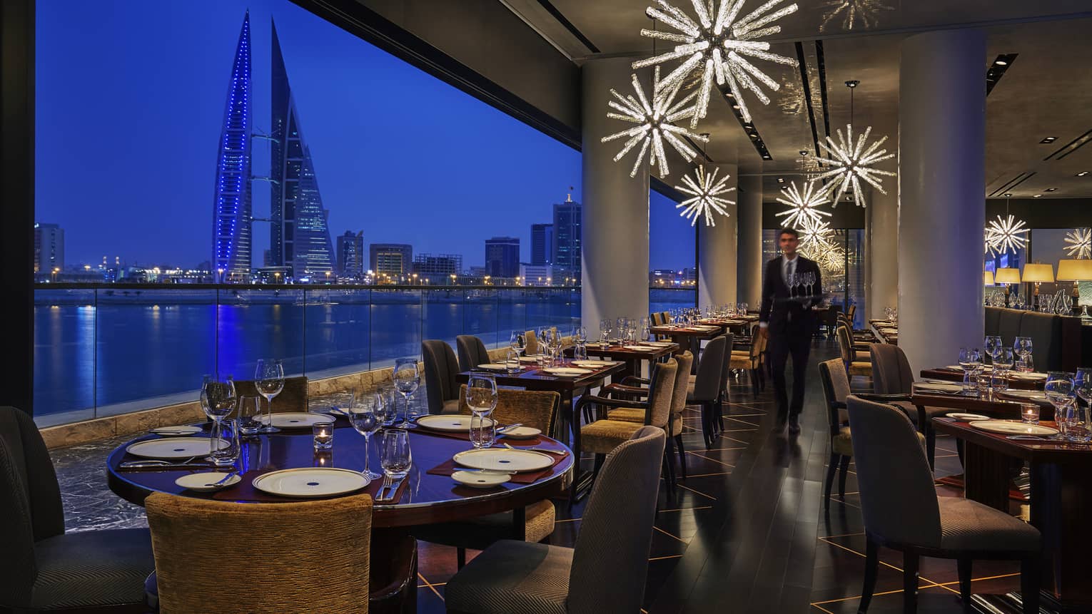 CUT by Wolfgang Puck dining room at night with formal tables, modern star-shaped chandeliers, gulf and modern tower views