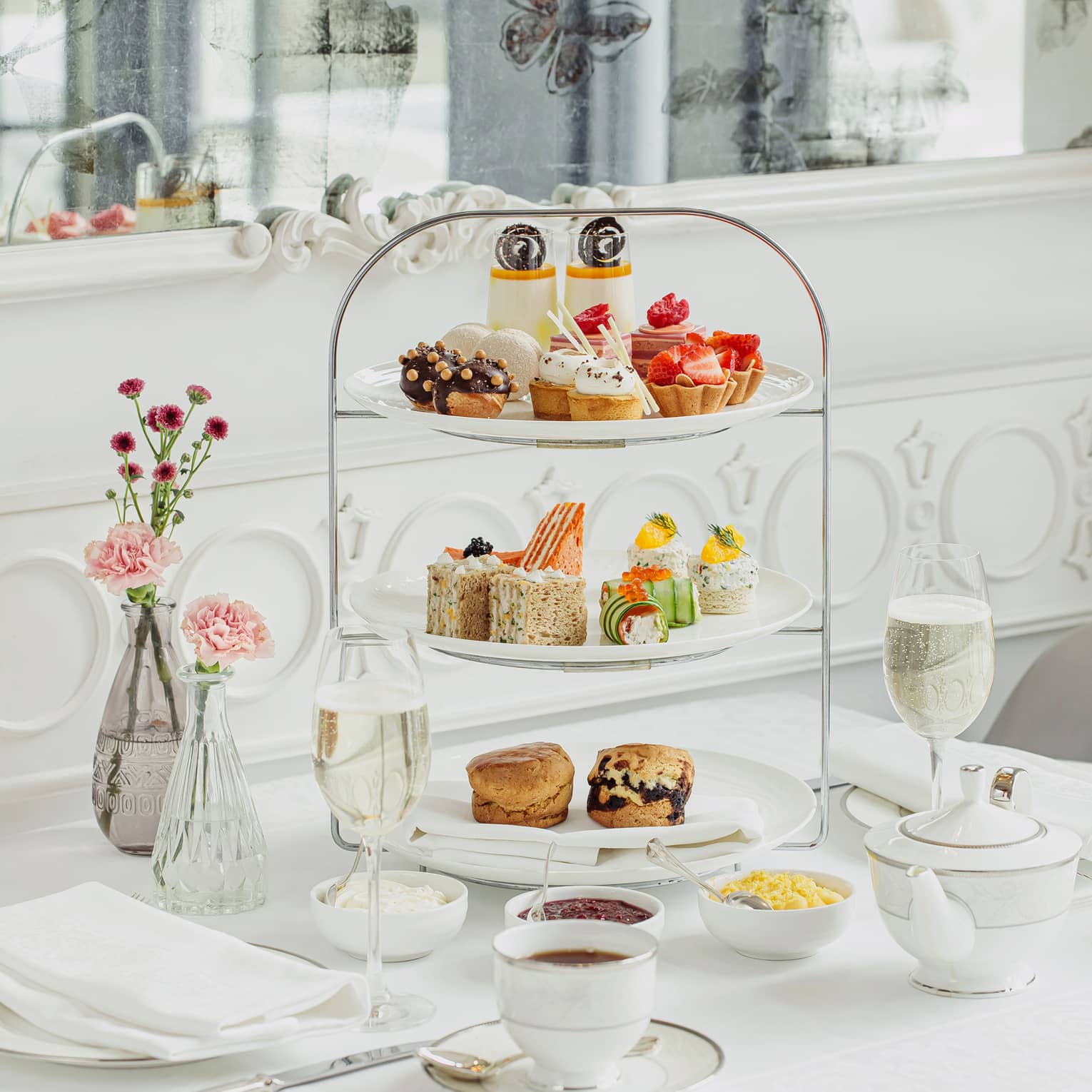 Side dishes, drinks and flowers surround a delicately tiered cake stand holding colourful selections of petite baked goods.