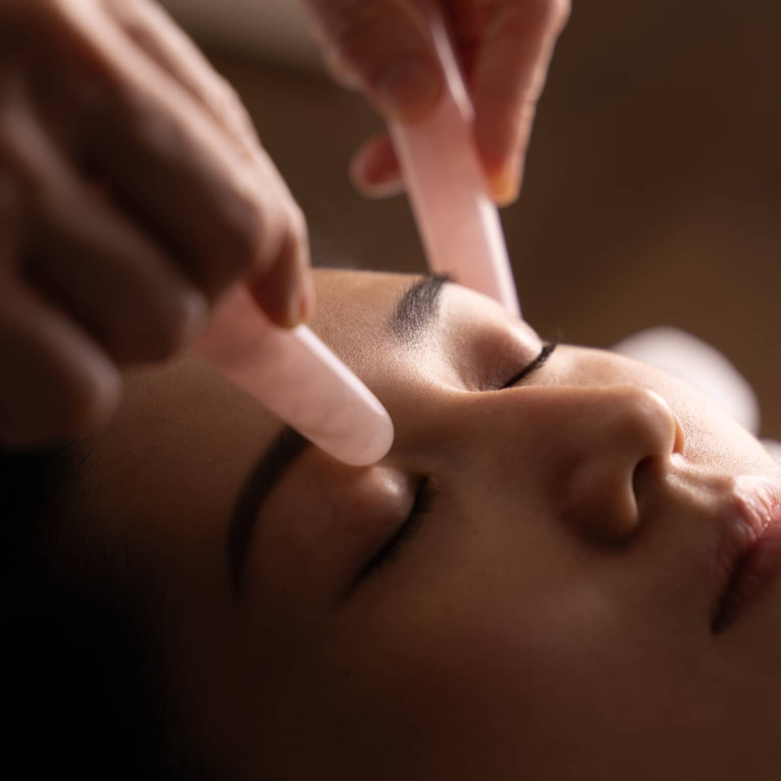 Hands hold pair of pink jade stones against woman’s eyelids during facial