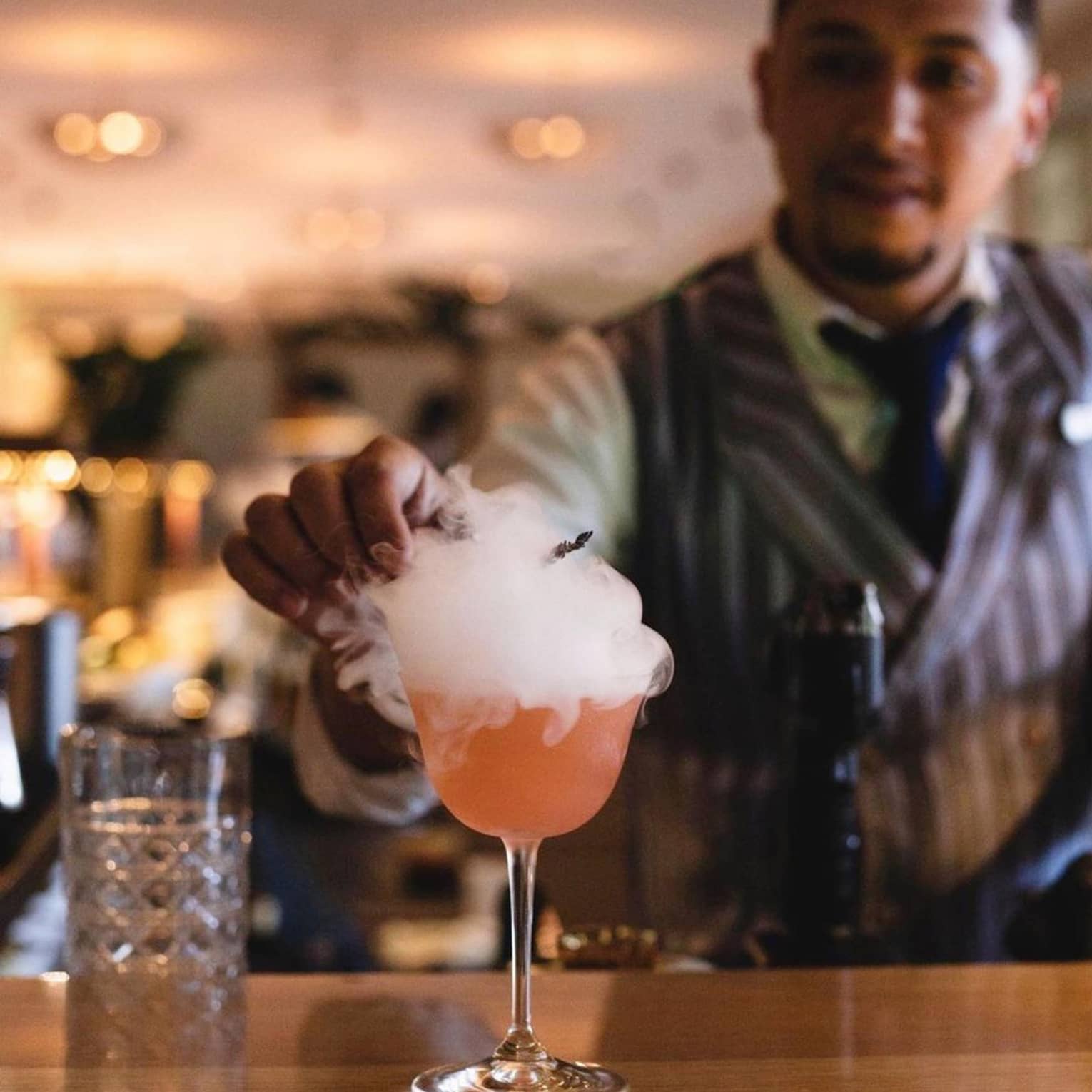 A mixologist garnishes a bright orange cocktail atop a smooth wooden bar; smoke billows over the sides of the stemmed glass.