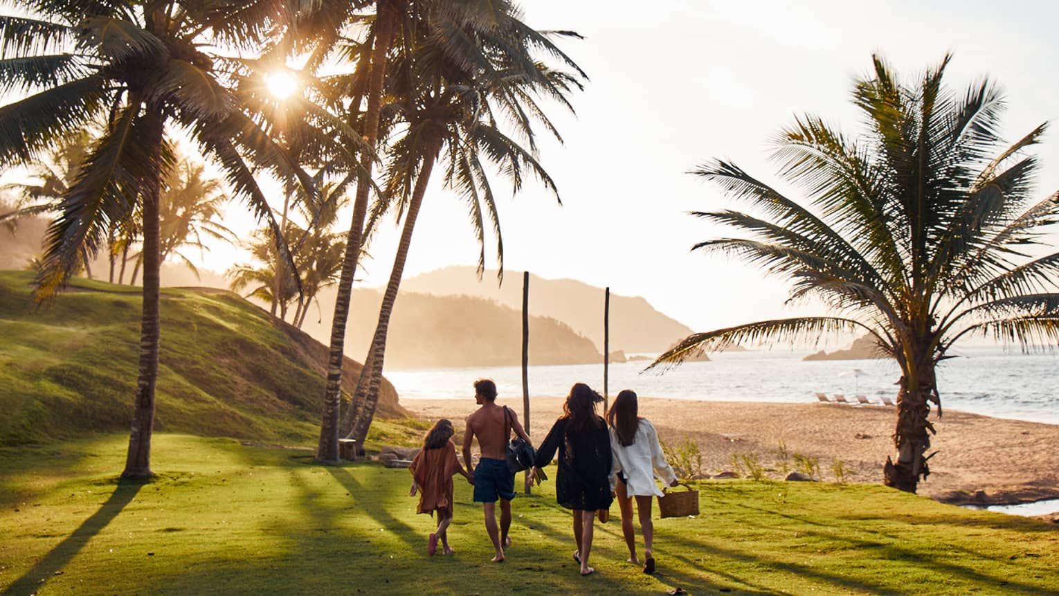 Rear view of a family walking toward a beach, one of them holding a picnic basket, as sunlight filters through a palm tree.