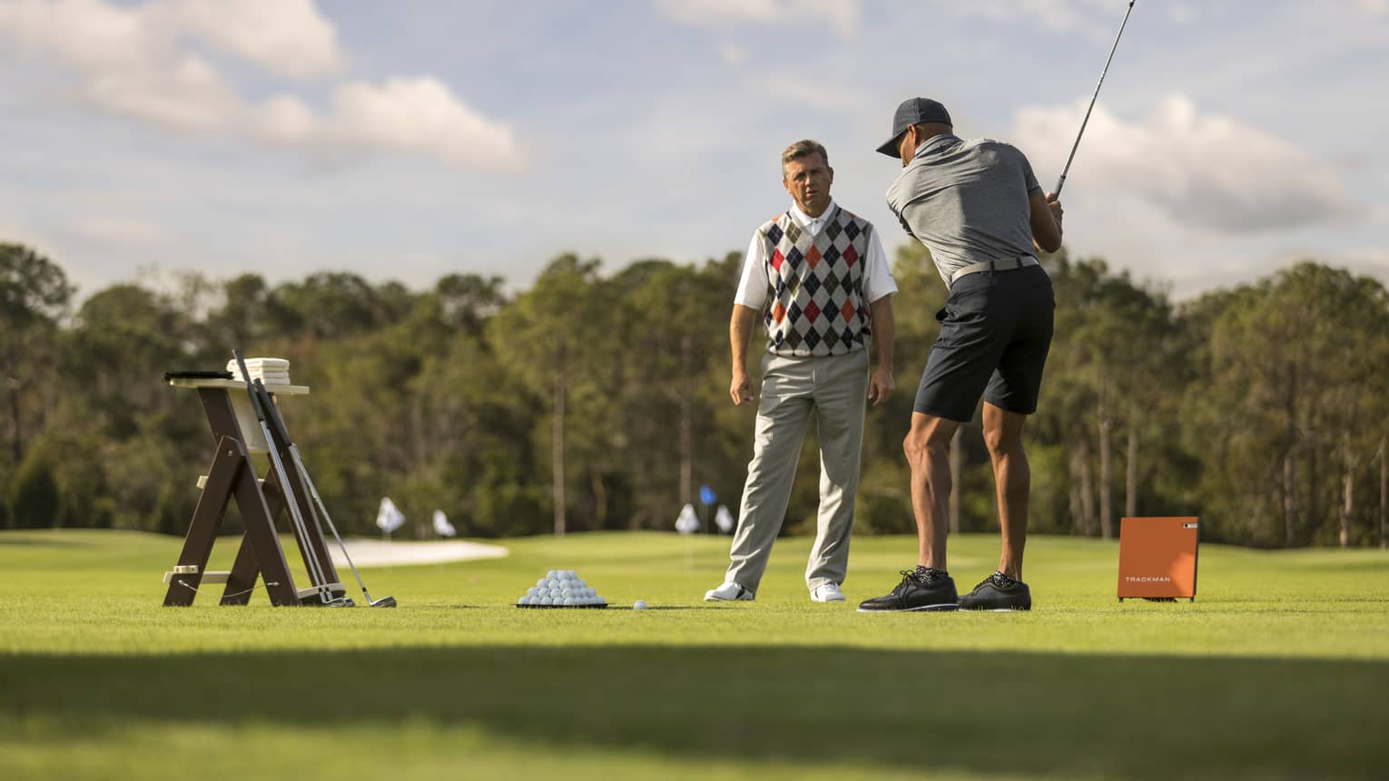Against clear sky, one golfer looks on as another prepares to swing, a tray of balls and a stand of clubs nearby.