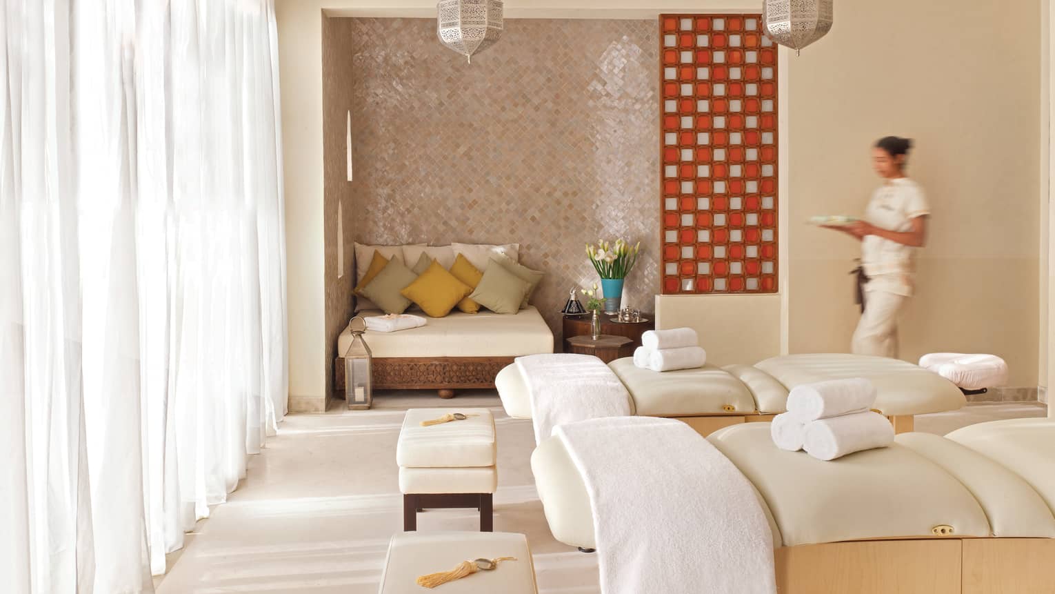 Spa staff walks into bright room by two massage beds with rolled white towels, window with long curtains
