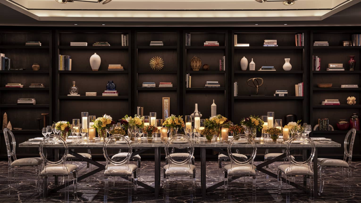 A large rectangular dining table with flowers and candles on top in a dimly lit room with books and items on shelves.