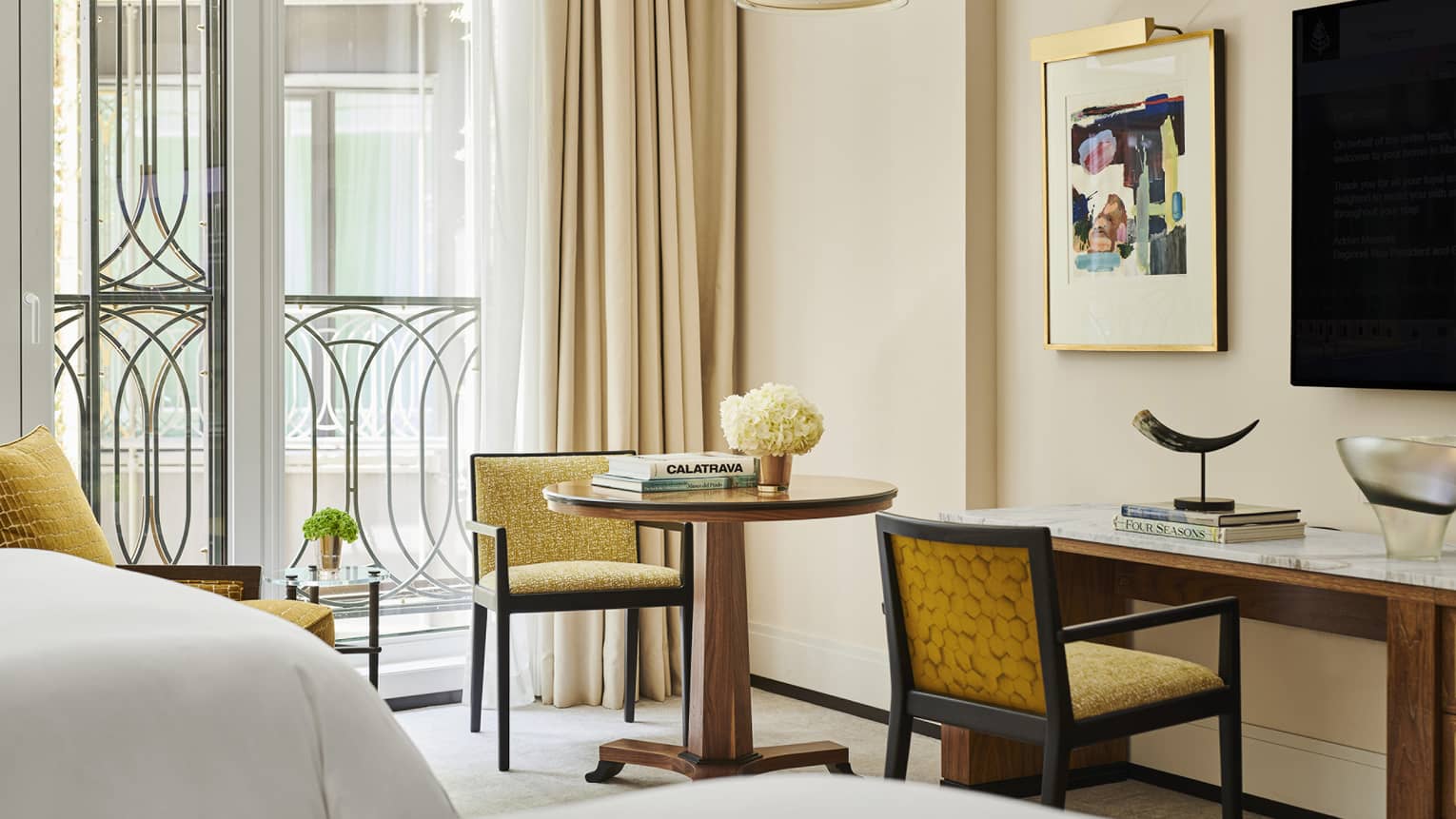 Hotel room seating area with round table, books, flowers, yellow chairs, abstract wall art, marble desk with horn sculpture and large windows with curtains