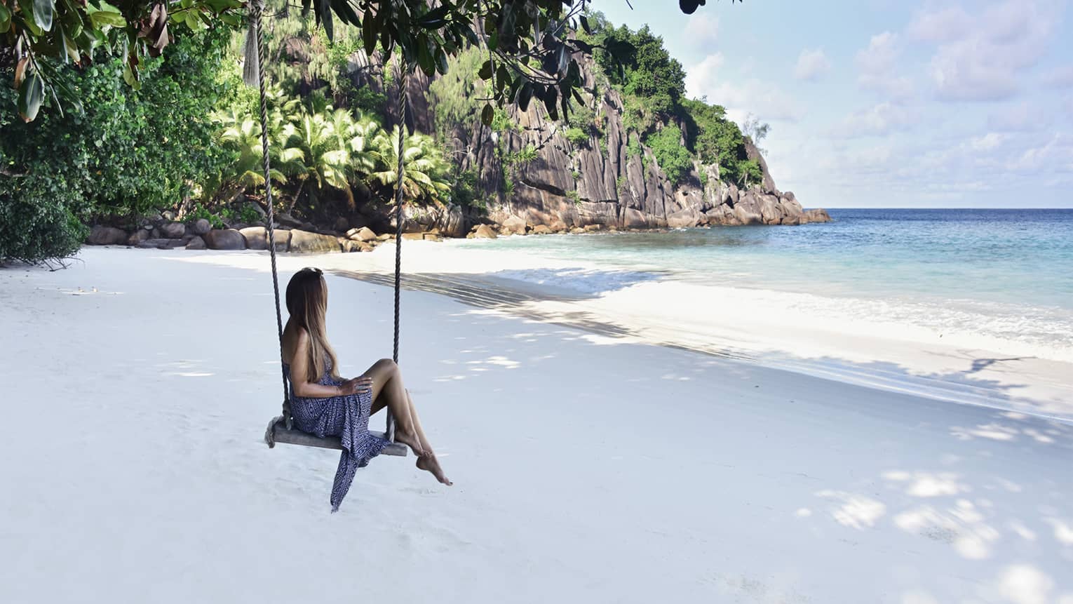 A woman sits on a tree swing on the beach with a view of a cliffside and the ocean