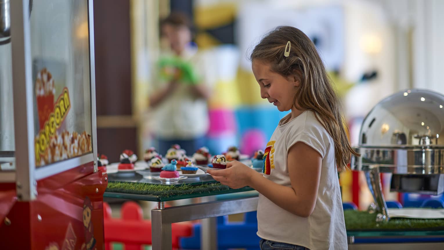 Young girl holds a cupcake at Friday Brunch, a carnival popcorn machine in front of her