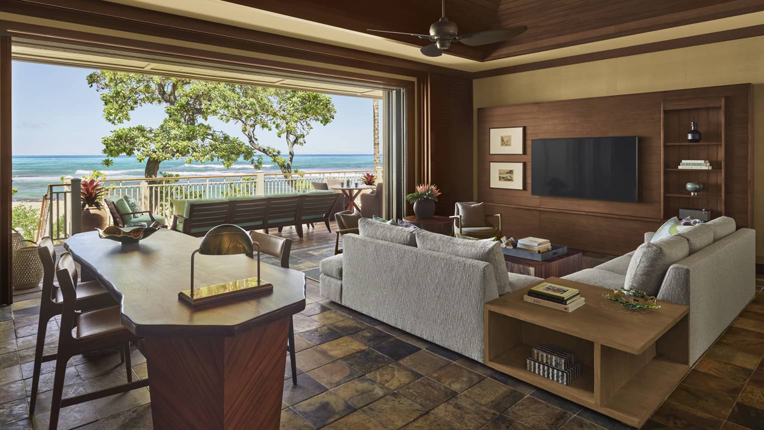 Living area with sectional grey sofa, wall TV, natural wood dining table, opening to beach terrace