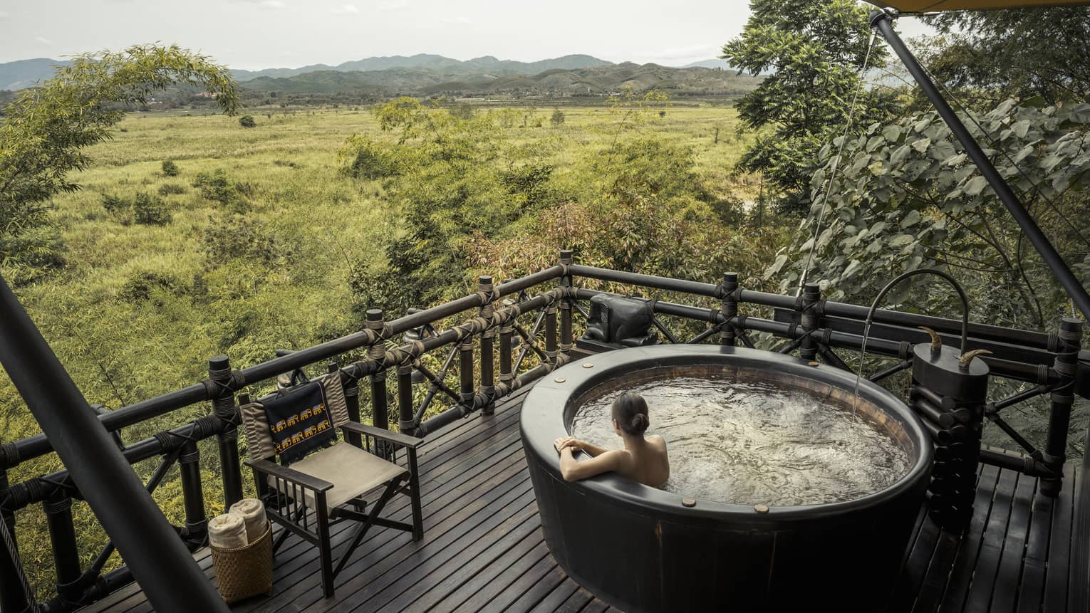 Woman wades in large round hot tub on wood deck overlooking green field, mountains