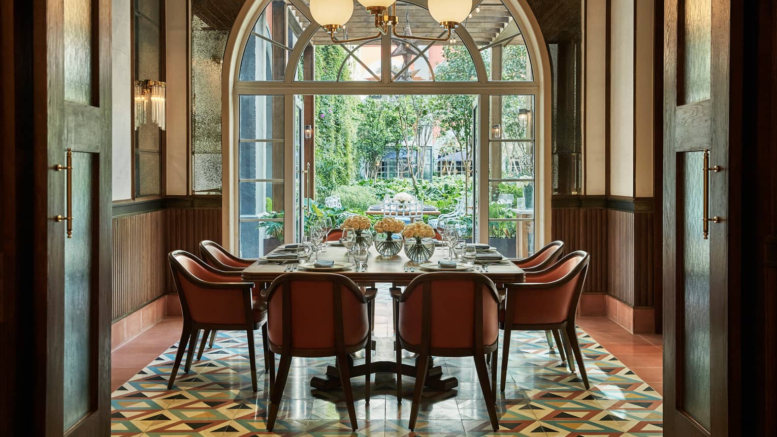 Dining table with orange chairs under globe lights by tall arched glass doors