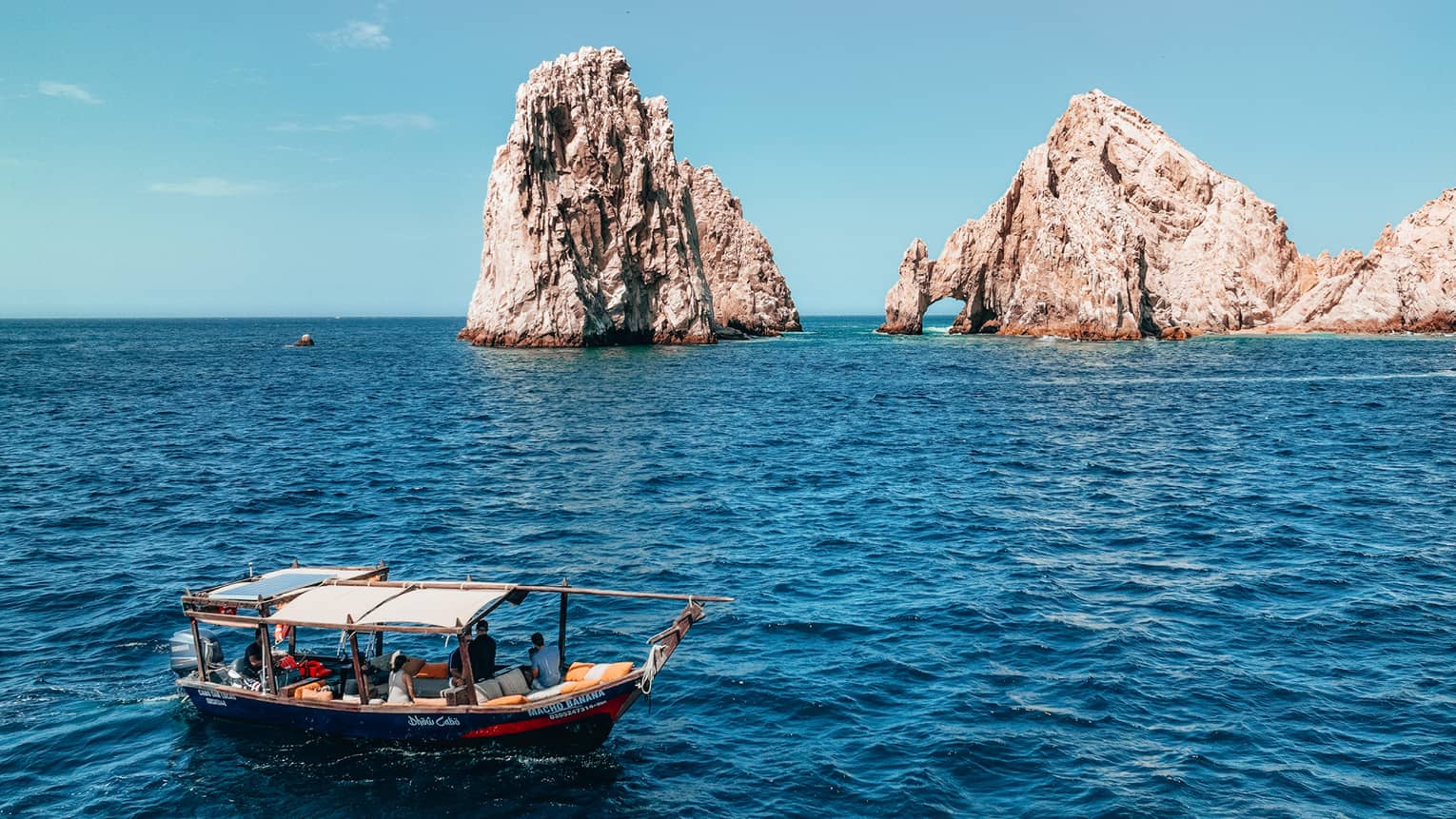 A canopied wooden tour boat in open water with tall craggy rock formations jutting out of the ocean in the background.