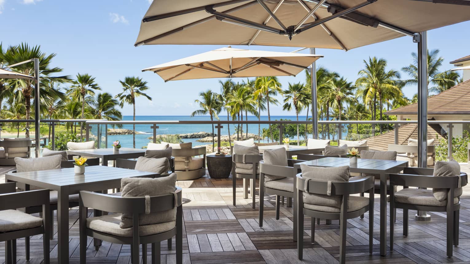 An outdoor dining space has wood deck floors, square tables shaded by large sun umbrellas and chairs with beige cushions and offers a view of palm trees and the ocean.