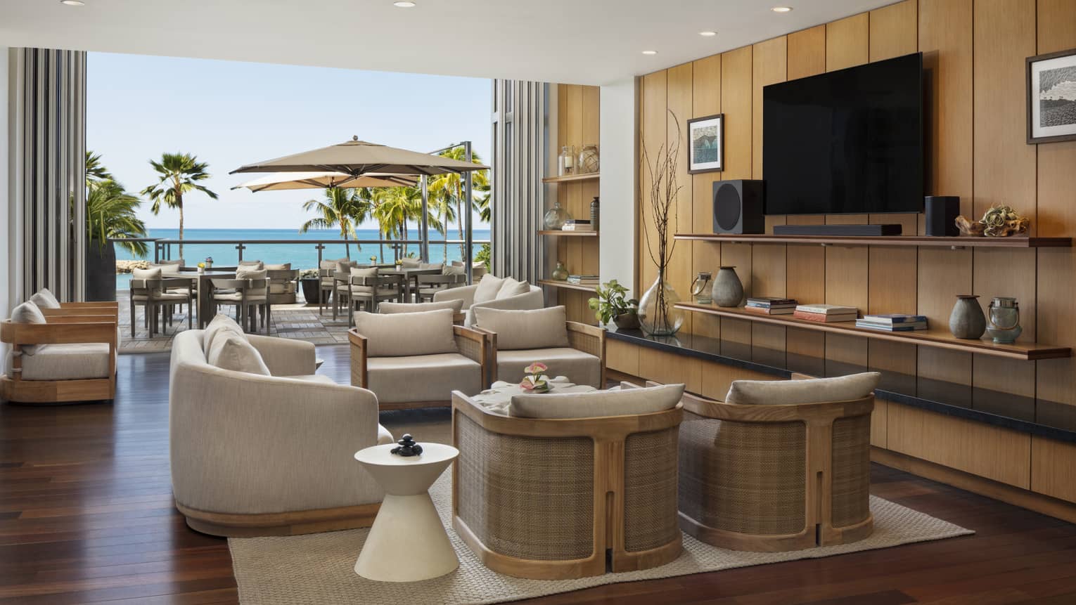 A well-lit interior space with dark wood floors, warm neutral walls and decor, a neutral area rug, and recessed ceiling lighting includes a floor-to-ceiling window with an ocean view, clusters of upholstered chairs, and decorative shelves and a flat-screen TV on one wall.